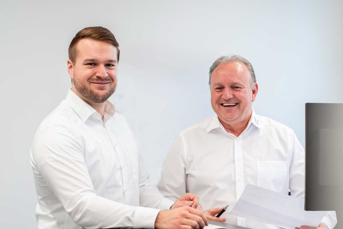 Zwei zufriedene Mitarbeiter im Büro, lächelnd mit Vertrag – attraktive Arbeitsbedingungen und Wertschätzung bei Elektro-Breitling GmbH